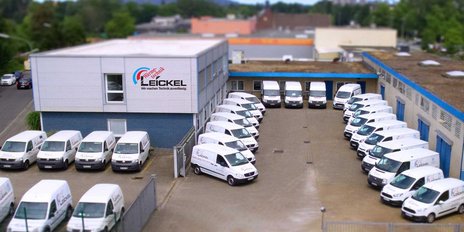 Eine Flotte von weißen Lieferwagen parkt ordentlich auf einem gepflasterten Hof vor einem modernen Gebäude.